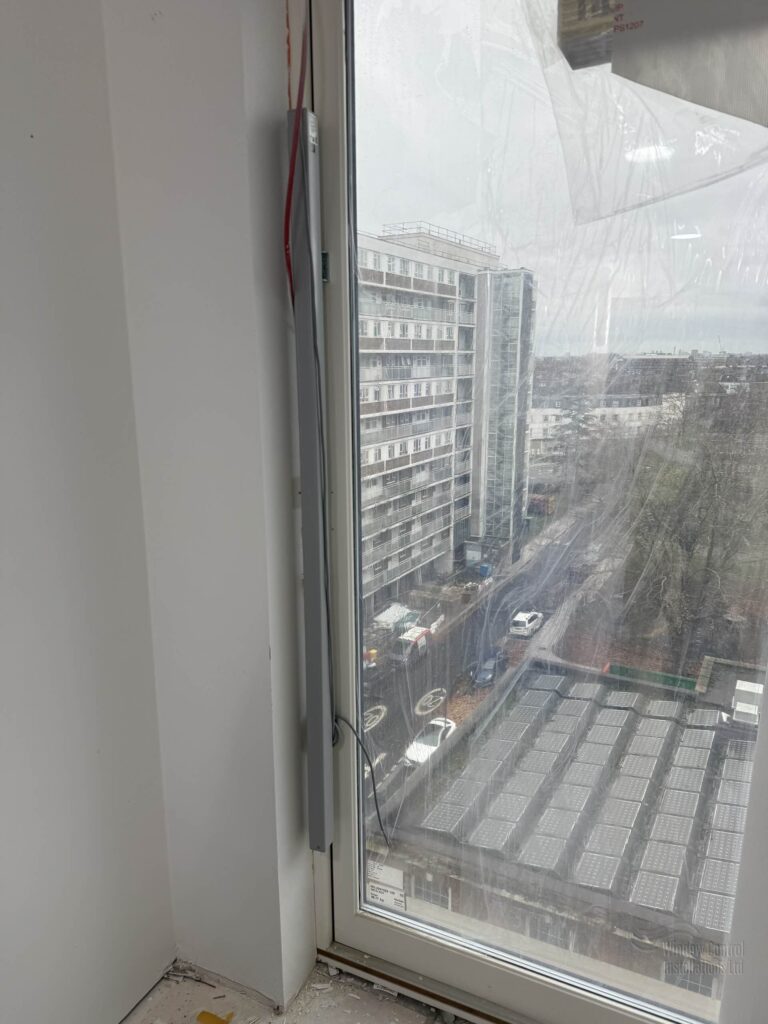 Close-up of a vertically mounted electric window actuator installed on a high-rise apartment block window.