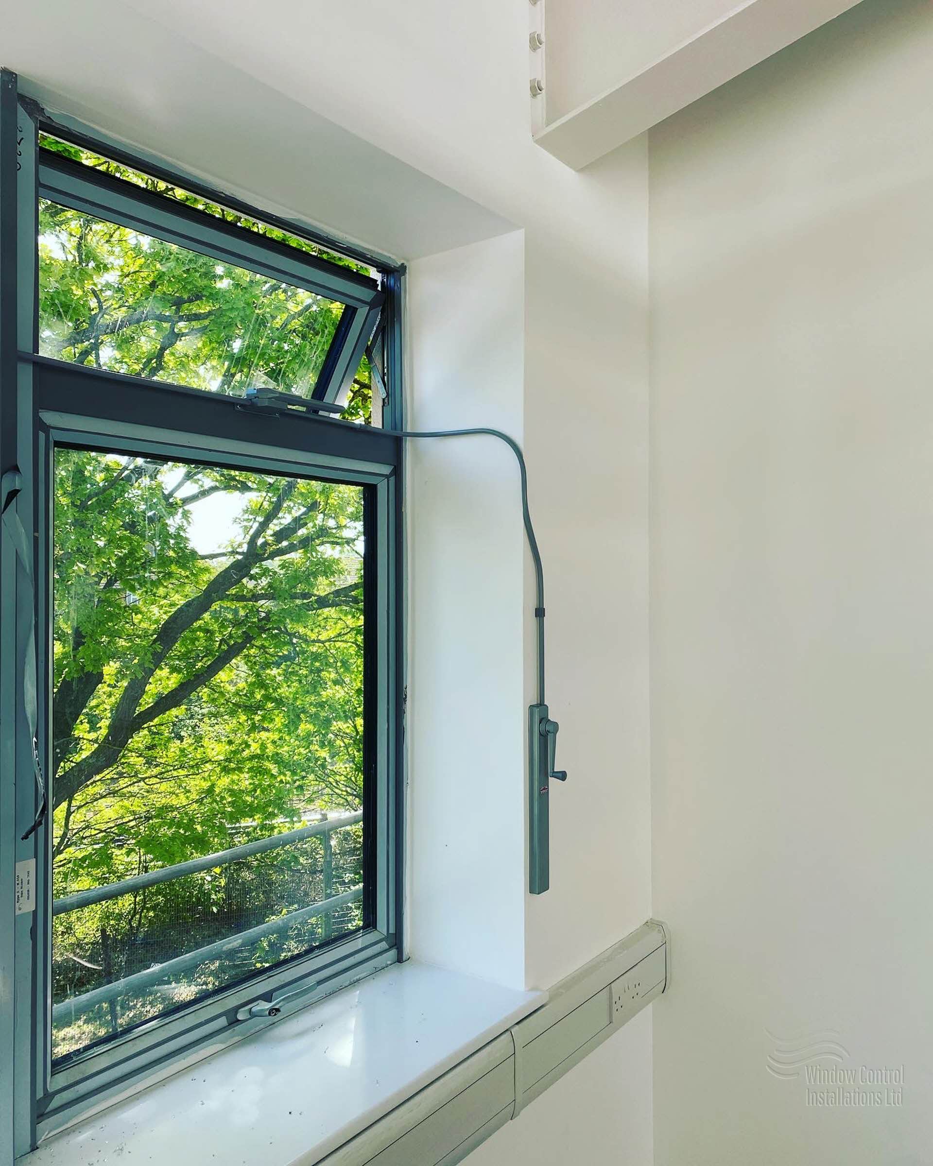 Teleflex manual window control system installed on a high-level commercial window, showing the chain opener, conduit, and wall-mounted operator handle.