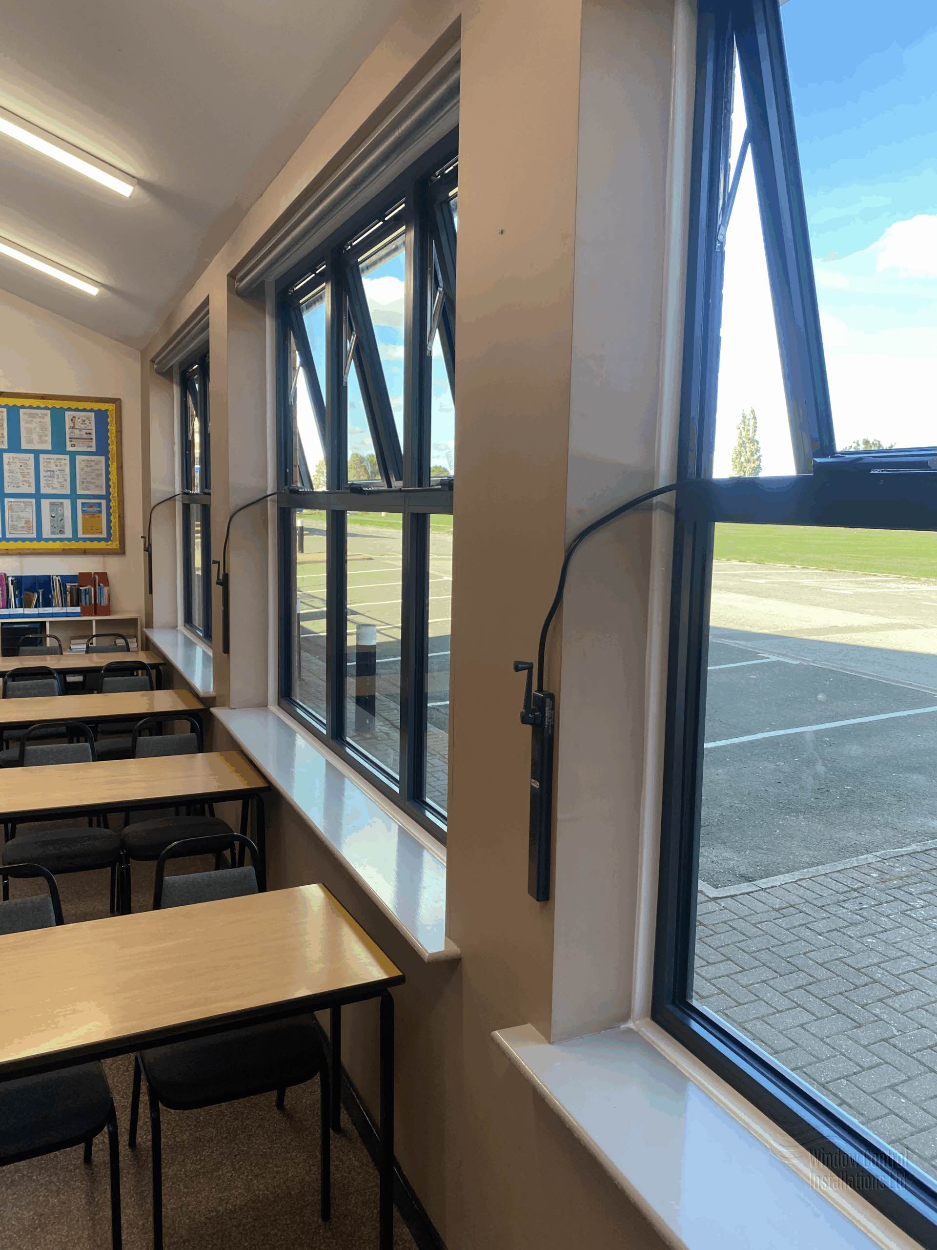 Teleflex manual window control system installed on dark grey aluminium top-hung windows in a commercial education setting.