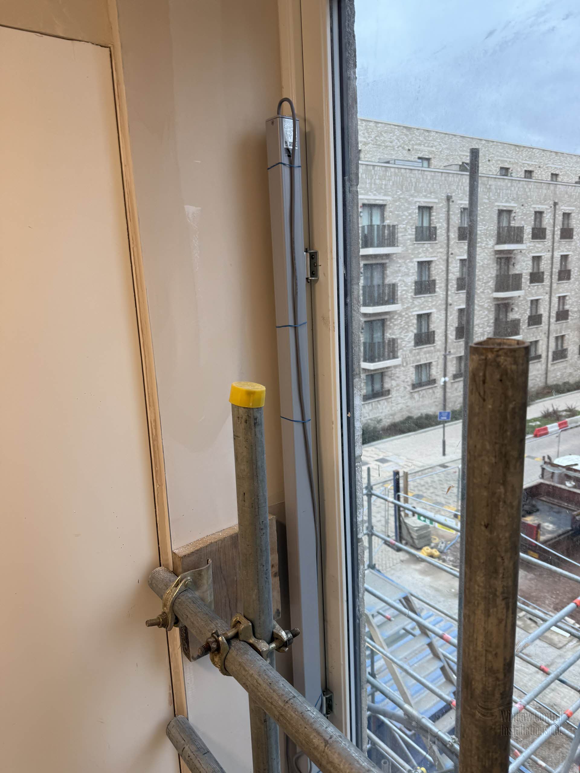 Vertical electric window actuator installed on a new build window frame, with construction scaffolding visible outside.